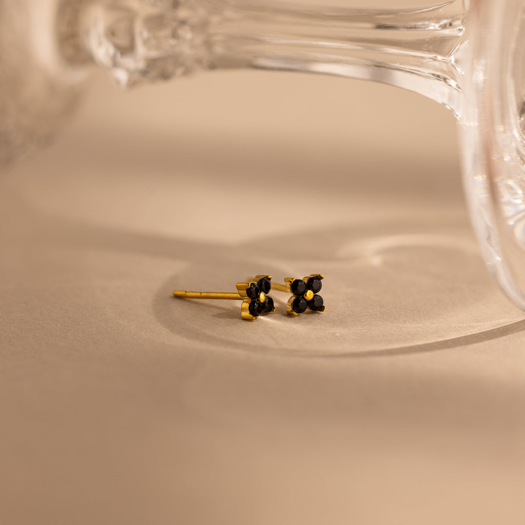 A pair of small gold stud earrings with black flower-like settings, photographed on a warm beige surface with a blurred clear glass object in the background.