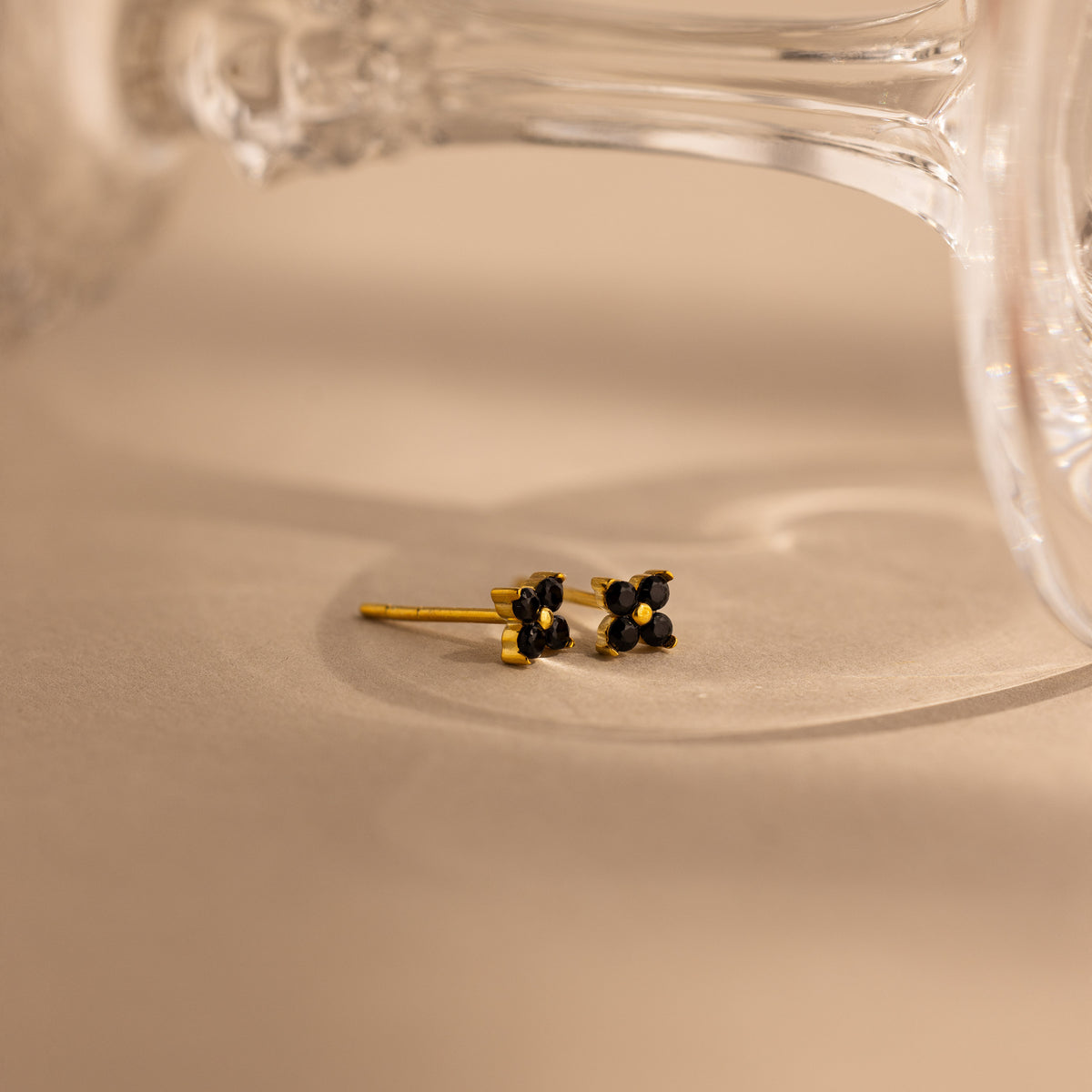 A pair of small gold stud earrings with black flower-like settings, photographed on a warm beige surface with a blurred clear glass object in the background.