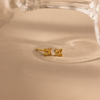A pair of small gold stud earrings with a butterfly-like design, resting on a beige surface with a soft-focus glass object in the background.