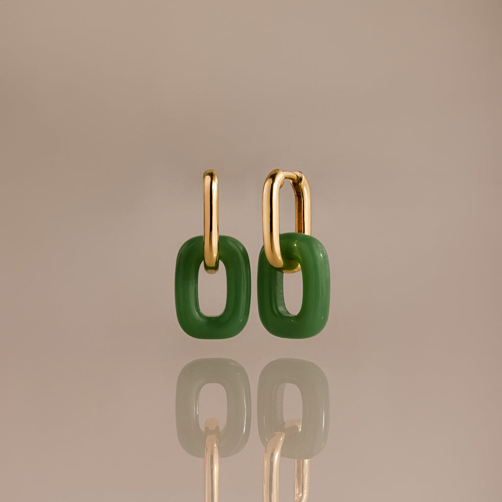 Gold hoop earrings with two connected rectangular jade links, reflecting on a surface.