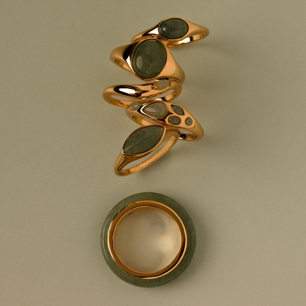 Five gold rings with green stones, including the Haven Jade Spinner Ring, are stacked above a round green and gold band bracelet on a beige background—an elegant display of mindful jewelry.