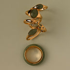 Five gold rings with green jade stones, stacked above a Jade Dewdrop Ring bracelet, radiate natural elegance on a beige background.