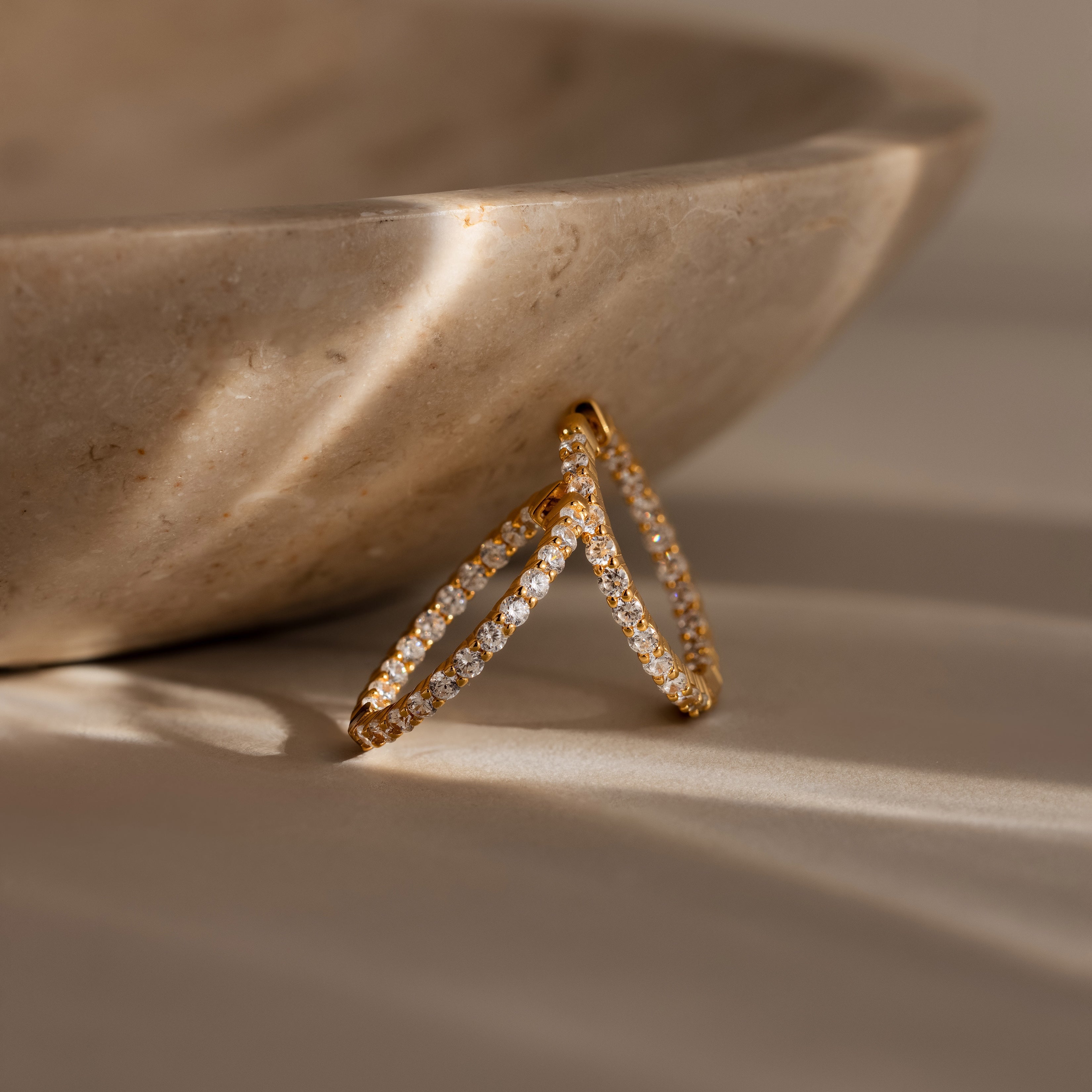 The Inside Out Diamond Hoops in 18K Gold, featuring sparkling diamonds and a timeless huggie design, are displayed against a beige stone bowl in gentle sunlight.