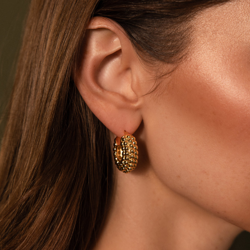 A close-up shows a woman wearing Beaded Dome Hoops, textured with gold beading. Her smooth brown hair and glowing skin highlight the earrings.