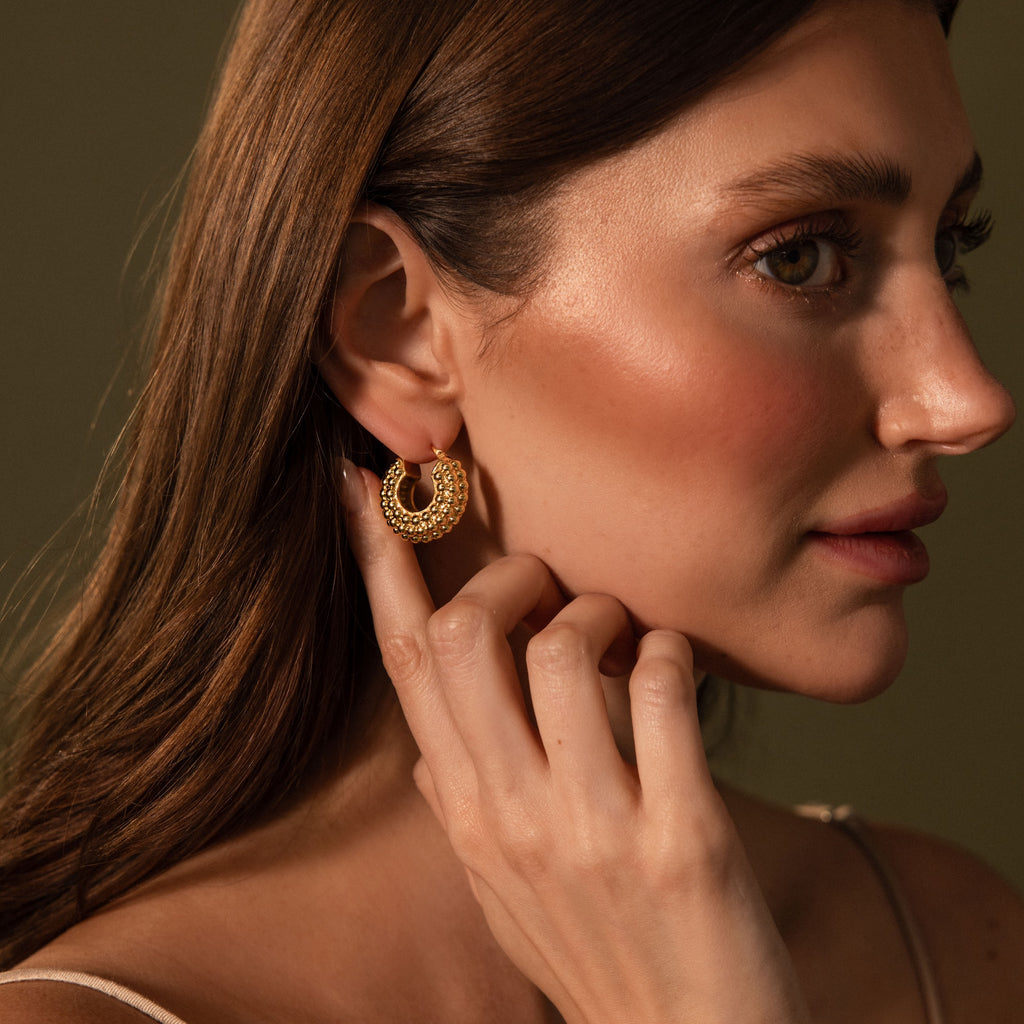 A woman with long brown hair touches Beaded Dome Hoops in 18K Gold, gazing to the side against a plain background.