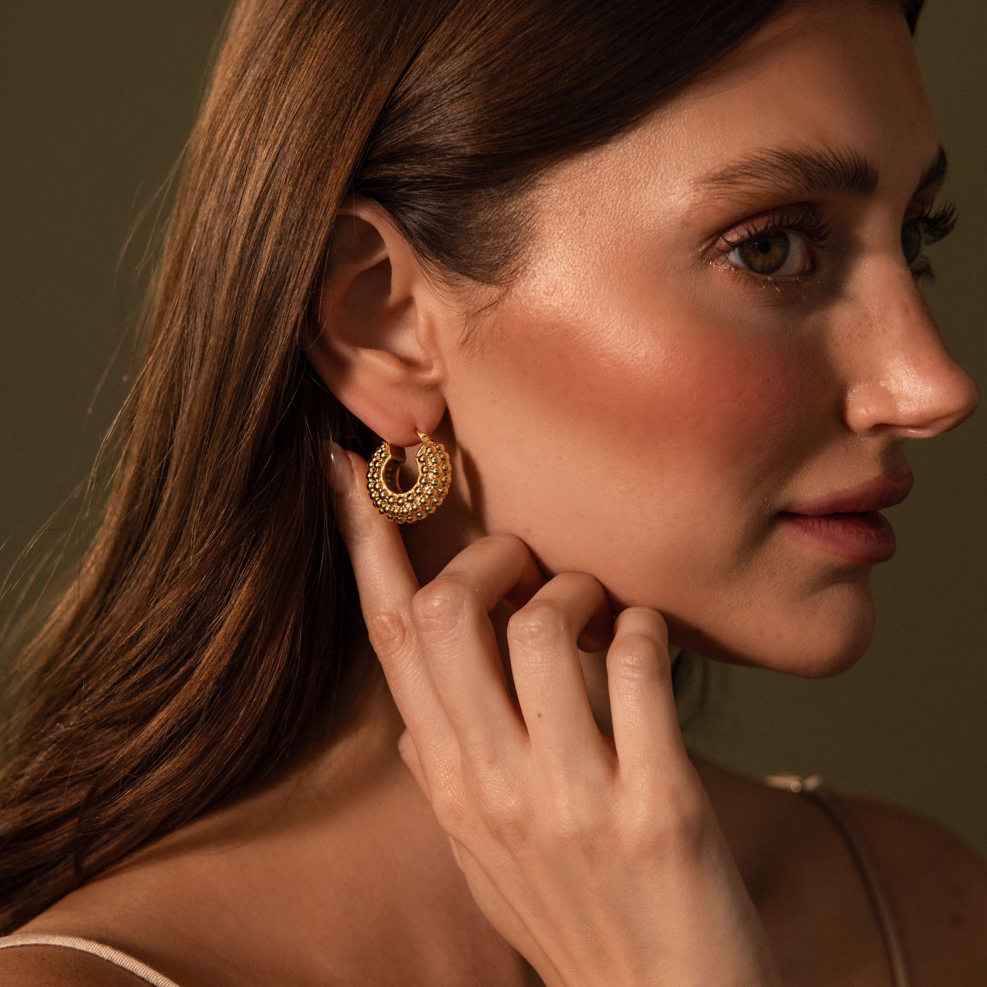 A woman with long brown hair touches Beaded Dome Hoops featuring gold beading against a neutral background.