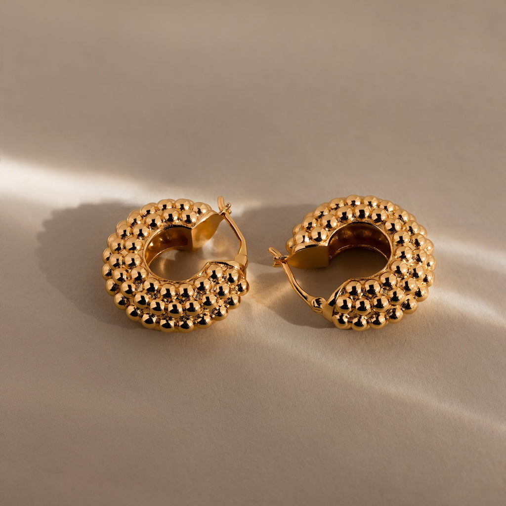 Beaded Dome Hoops in 18K Gold rest on a neutral surface, softly lit to highlight their textured gold beading and bold statement design.