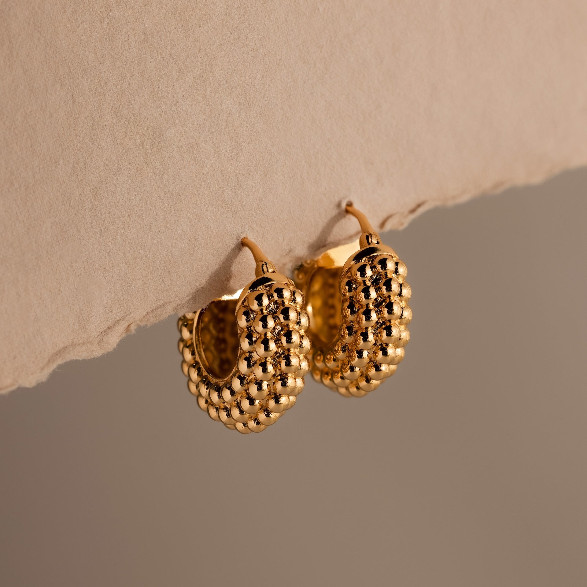 The Beaded Dome Hoops in 18K Gold feature intricate gold beading and are displayed elegantly against a neutral beige background for a refined touch.