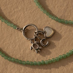 Beaded Jade Clasp Necklace in Silver features green jade beads, silver letter C, heart, and crab charms, plus a gold clasp—ideal for layering. Shown on a beige textured background.