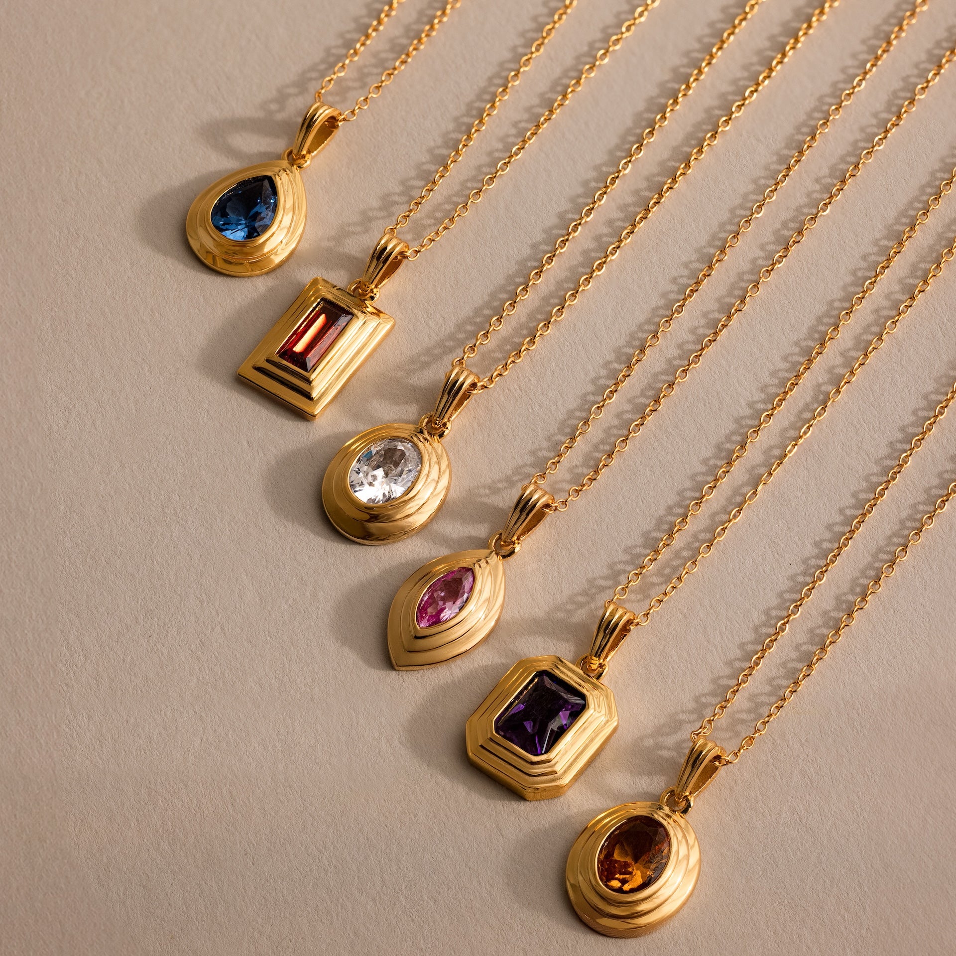 Five vintage-inspired gold necklaces, including the Ribbed Birthstone Pendant Necklace, each feature a unique colored gemstone pendant and are arranged neatly on a beige surface.