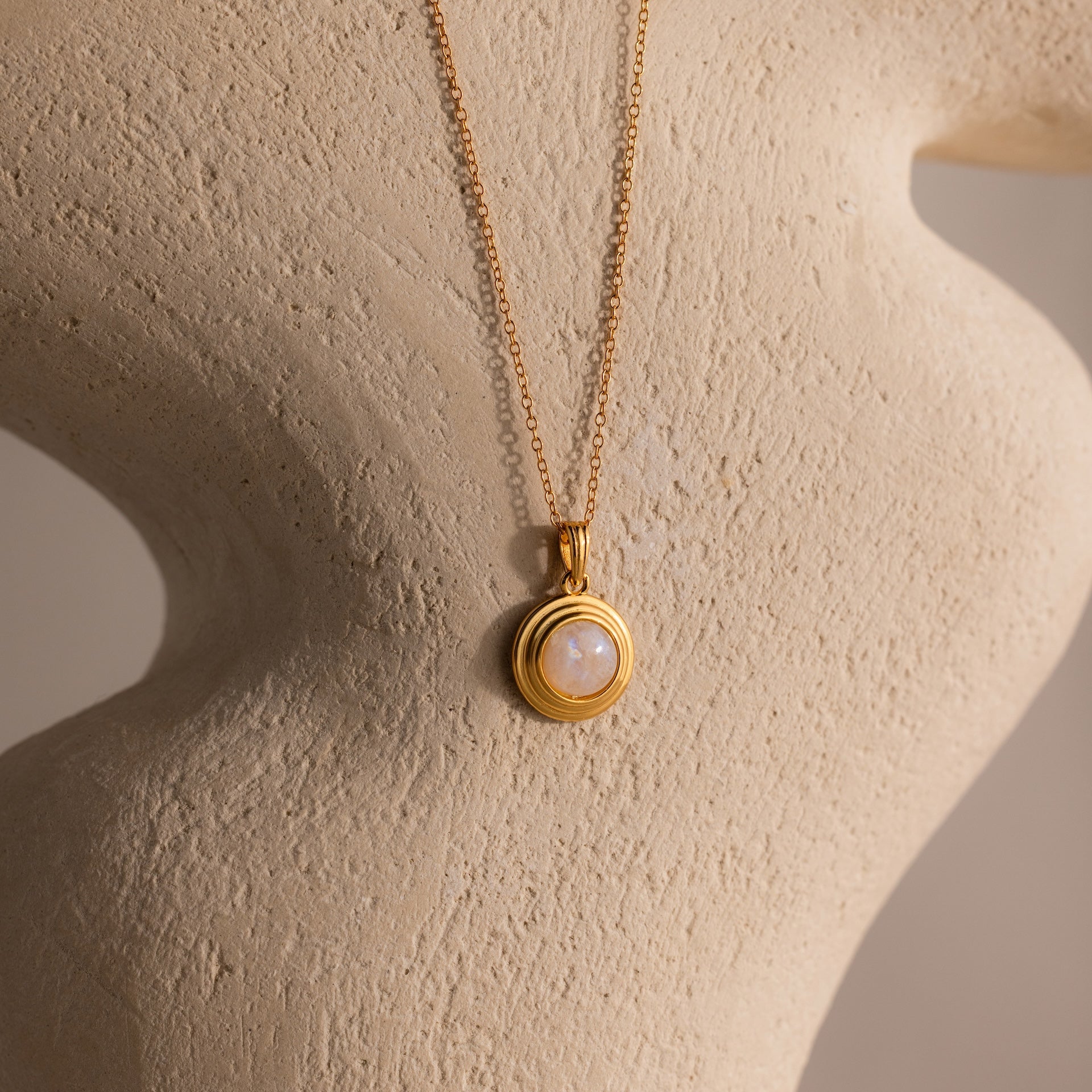 Ribbed Moonstone Pendant Necklace featuring a gold ribbed chain and a round white moonstone pendant, displayed on a textured beige sculpture.