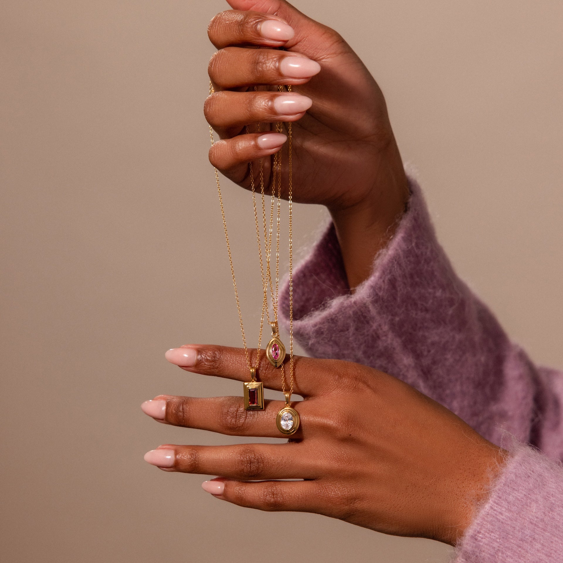 A hand in a purple sweater holds three gold necklaces, including a Birthstone Pendant Necklace and the Ribbed Diamond Pendant Necklace, each featuring vintage-inspired accents, set against a beige background.