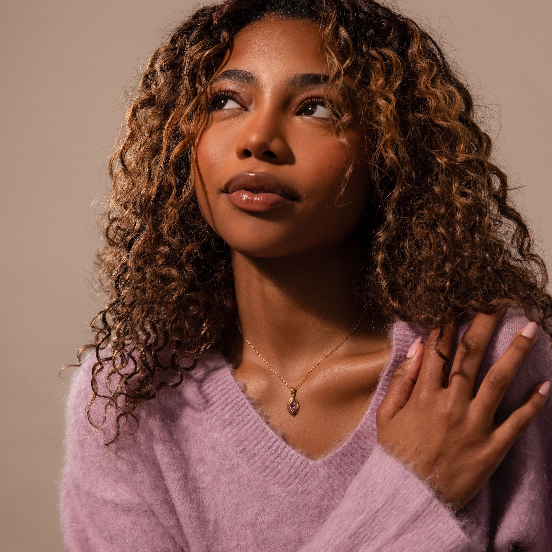 Woman with curly hair in a soft purple sweater, gazing upward with hand on shoulder, wears the Ribbed Birthstone Pendant Necklace—a delicate gold accessory—against a neutral background.