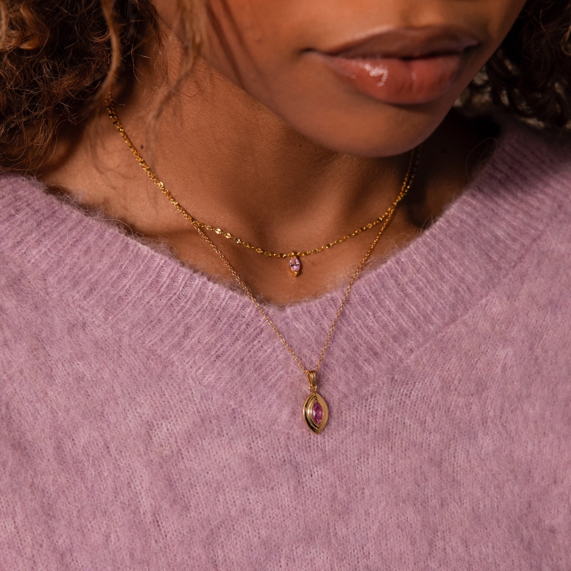 Woman wearing a lavender sweater and two gold necklaces, including the Ribbed Alexandrite Pendant Necklace featuring a ribbed gold chain and pink gemstone pendant.
