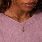 A woman wears a Ribbed Emerald Pendant Necklace layered with a vintage gemstone necklace, both showcasing purple pendants, and pairs them with a soft lavender sweater.