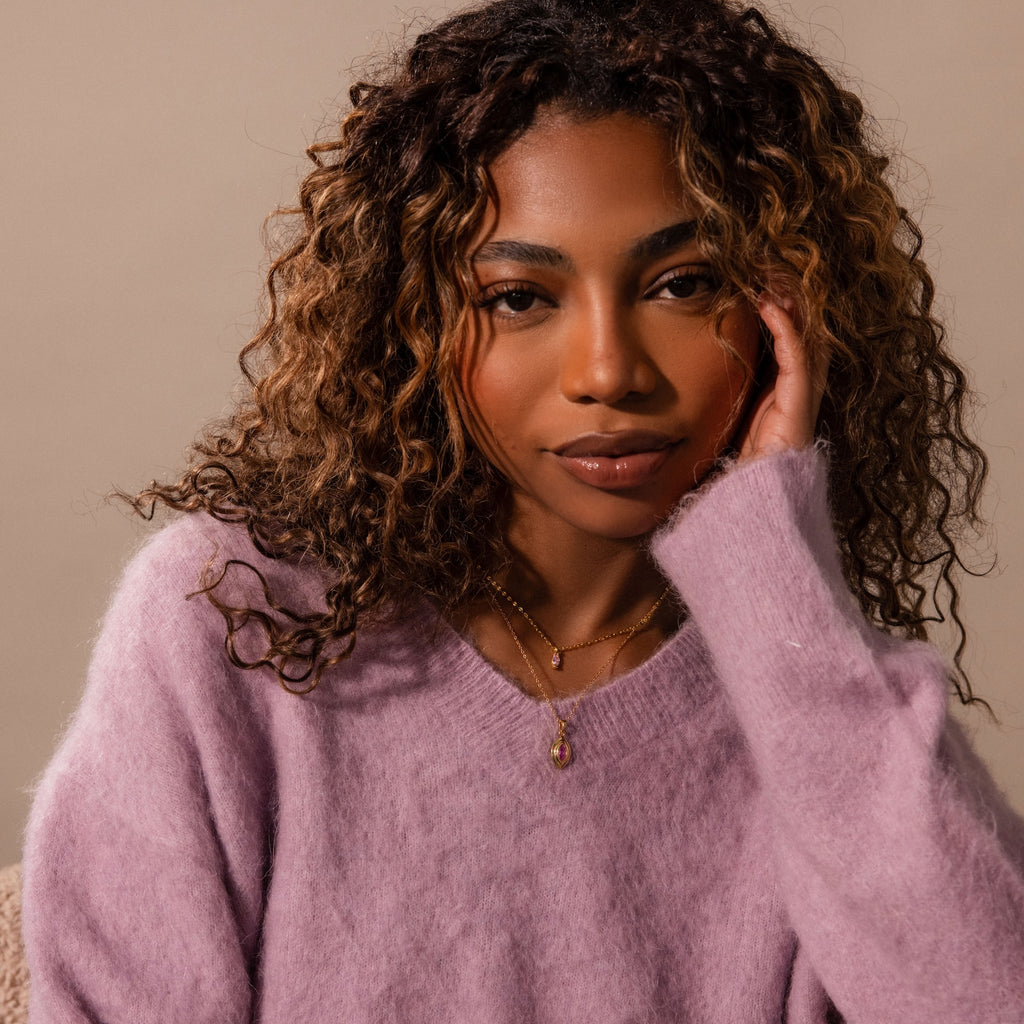 Woman with curly hair wearing a fuzzy lavender sweater and vintage-inspired jewelry, including the Ribbed Birthstone Pendant Necklace, posing with her hand on her face.