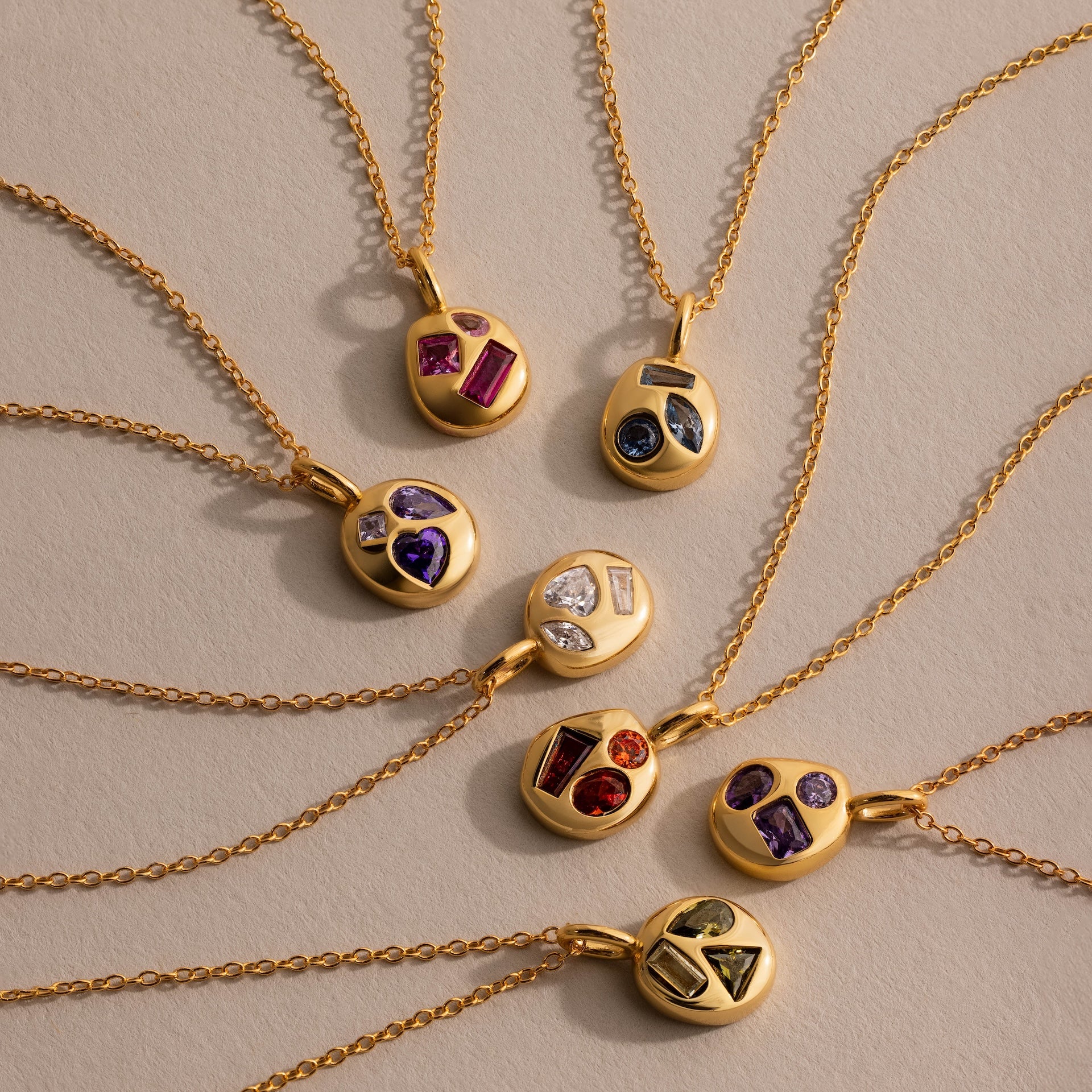 Eight gold necklaces, including the Mixed Aquamarine Capsule Pendant Necklace, showcase vibrant gemstone pendants arranged in a circular pattern on a beige surface.