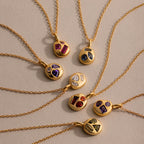 Seven gold birthstone necklaces with colorful gemstone pendants, featuring the Mixed Emerald Capsule Pendant Necklace, are arranged in a circle on a beige surface.