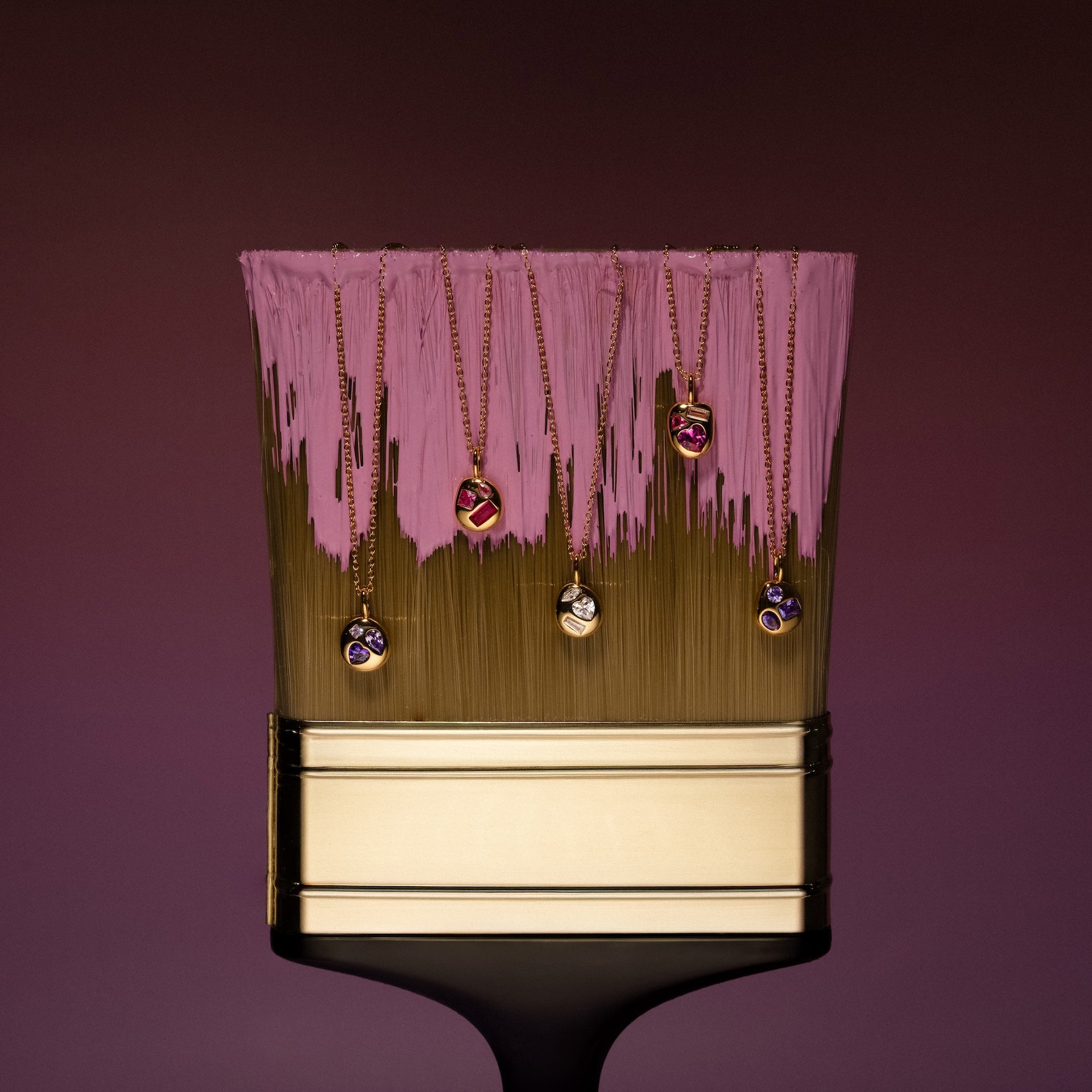The Mixed Pink Tourmaline Capsule Necklace shines with vibrant gemstones in a gold setting, displayed on a paintbrush dipped in pink paint against a gradient backdrop.