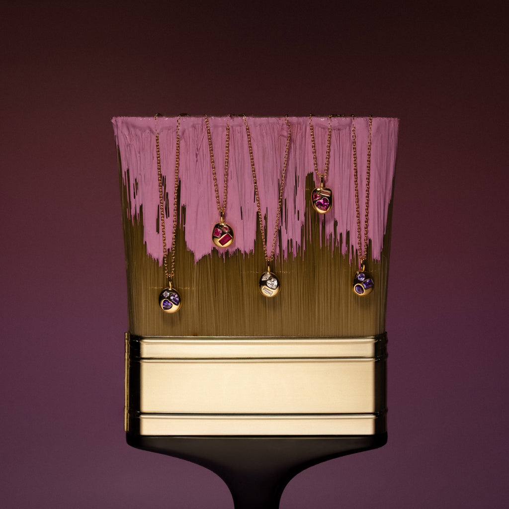 A paintbrush with pink paint displays six gold necklaces, featuring the Mixed Ruby Capsule Pendant Necklace with colorful gemstone pendants hanging from its bristles.