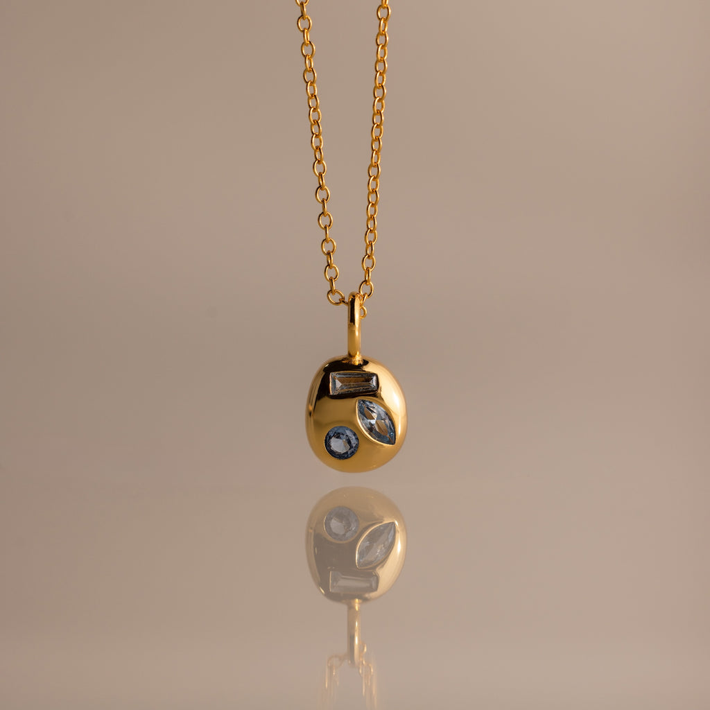 Mixed Aquamarine Capsule Pendant Necklace featuring a round charm with three different shapes of aquamarine gemstones, displayed on a beige background.