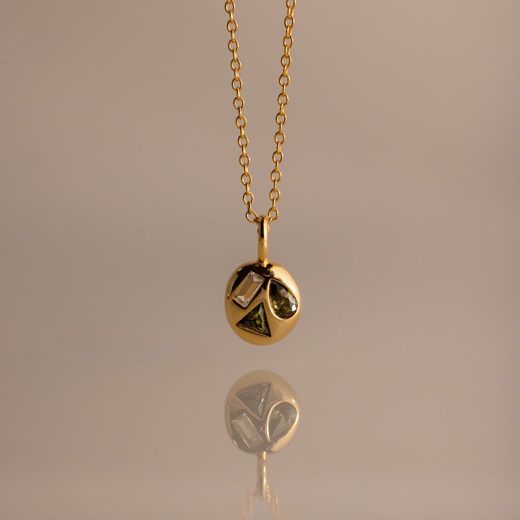 Mixed Peridot Capsule Pendant Necklace with a round pendant featuring geometric shapes, reflected on a smooth surface.