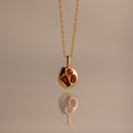 Mixed Garnet Capsule Pendant Necklace with a round pendant featuring three red gemstones, reflected on a smooth surface.