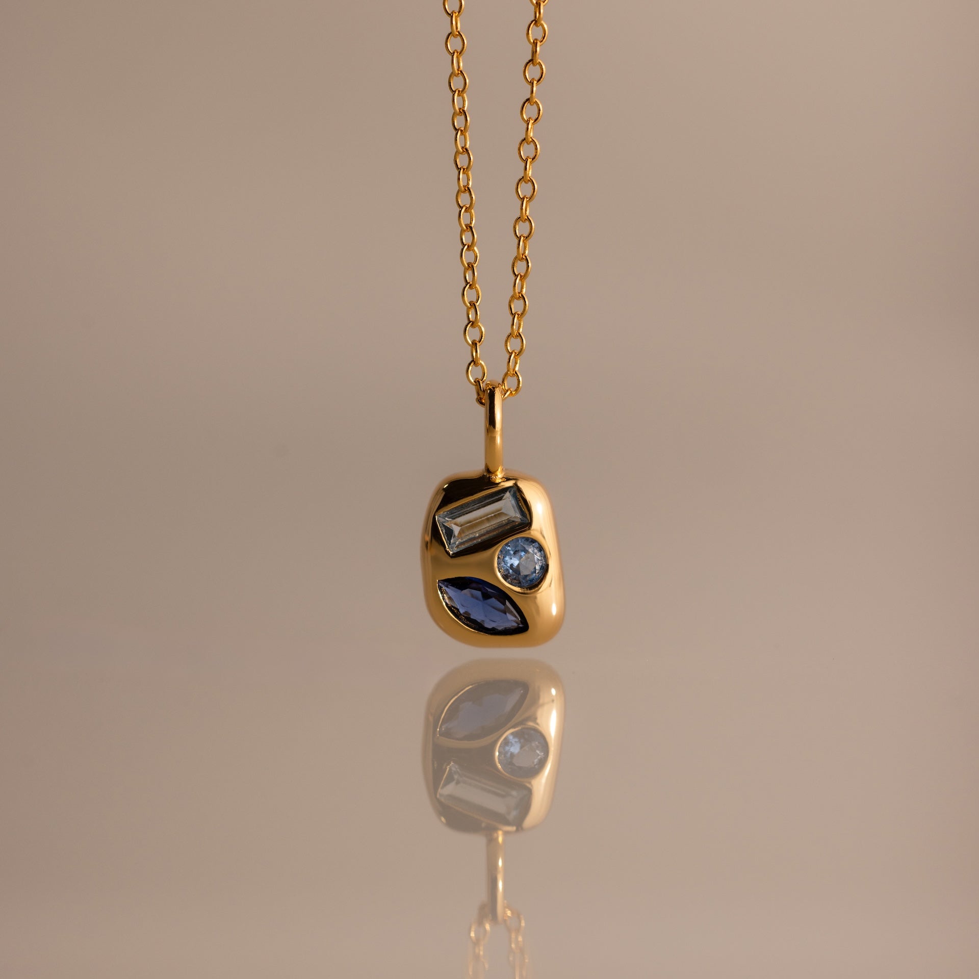 Mixed Blue Zircon Capsule Pendant Necklace, featuring three uniquely shaped blue zircons in a capsule pendant, reflected on a smooth surface.