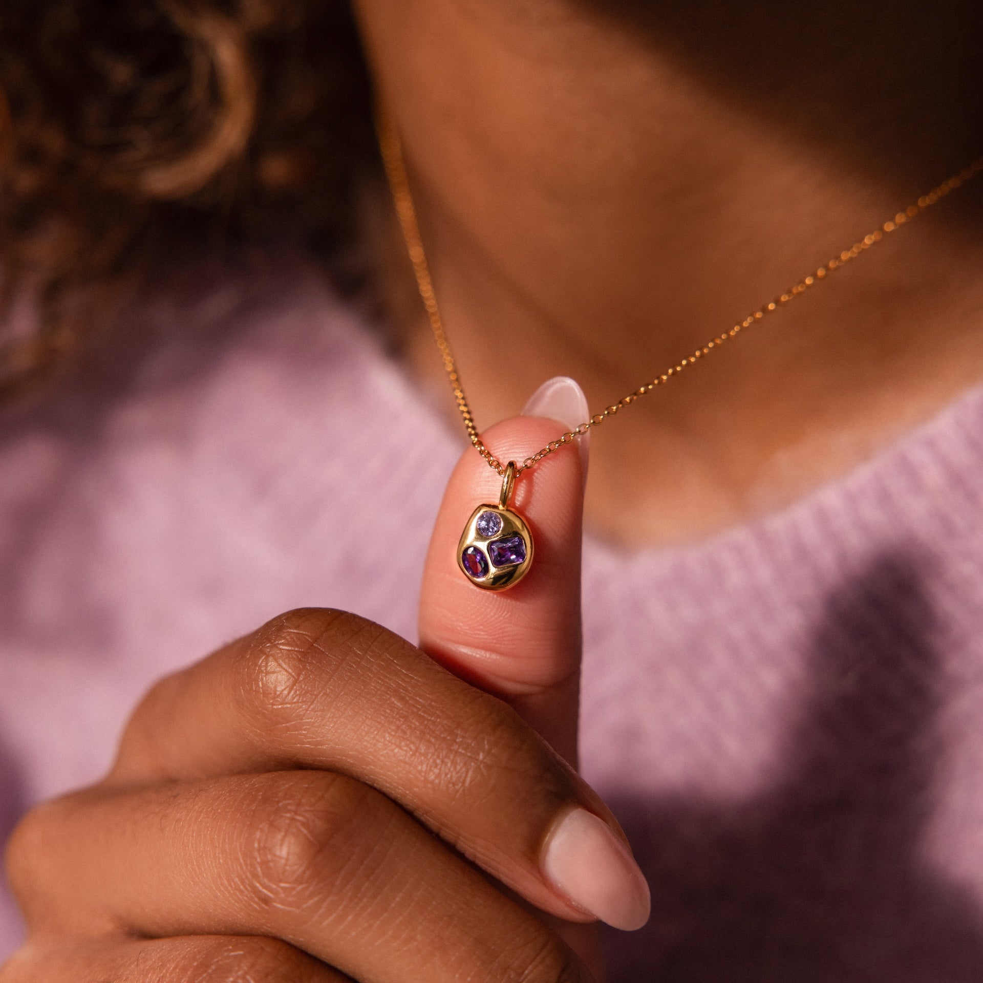 A hand holds the Mixed Amethyst Capsule Pendant Necklace, featuring a gemstone pendant with purple and red stones, styled over a lilac sweater.