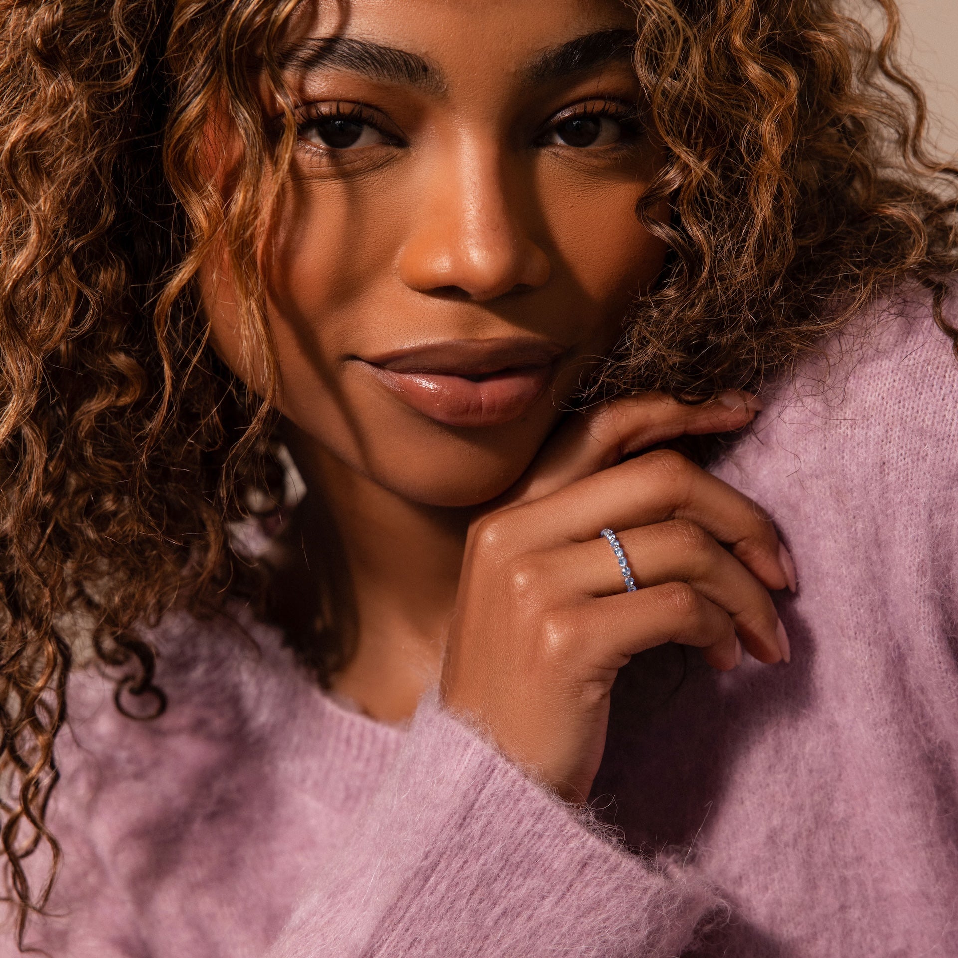 A woman with curly hair, dressed in a purple sweater, smiles softly while showcasing the Lace Eternity Blue Zircon Ring on her finger.