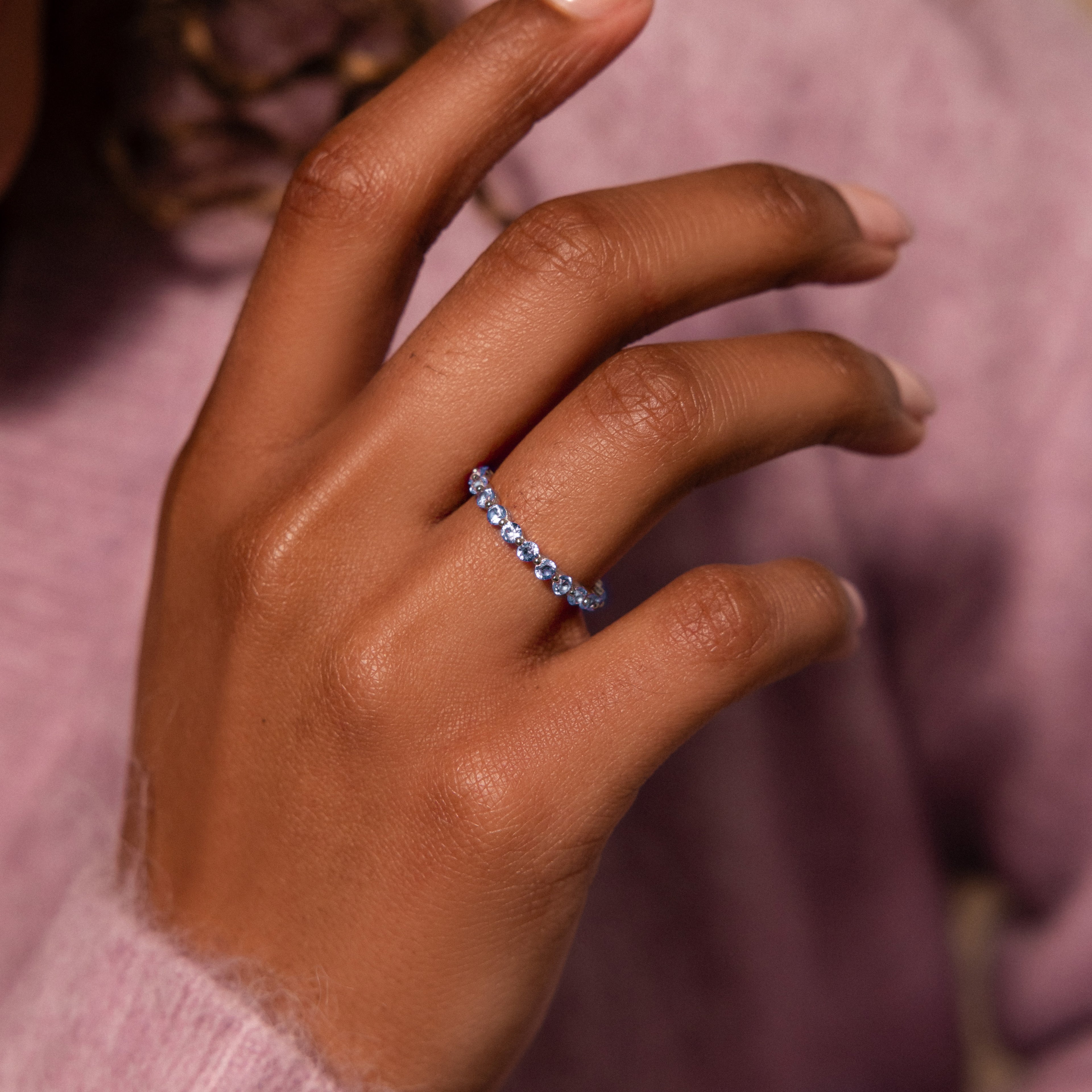 A hand wearing a delicate Lace Eternity Peridot Ring with sparkling green peridot gemstones, set against a soft purple sweater background.