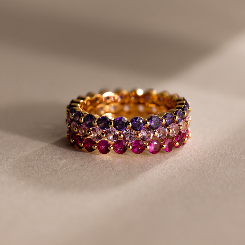 The Lace Eternity Ruby Ring, featuring three stacked gold bands with purple, clear, and pink gemstones, creates an elegant birthstone display against a neutral background.