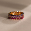 The Lace Eternity Ruby Ring, featuring three stacked gold bands with purple, clear, and pink gemstones, creates an elegant birthstone display against a neutral background.
