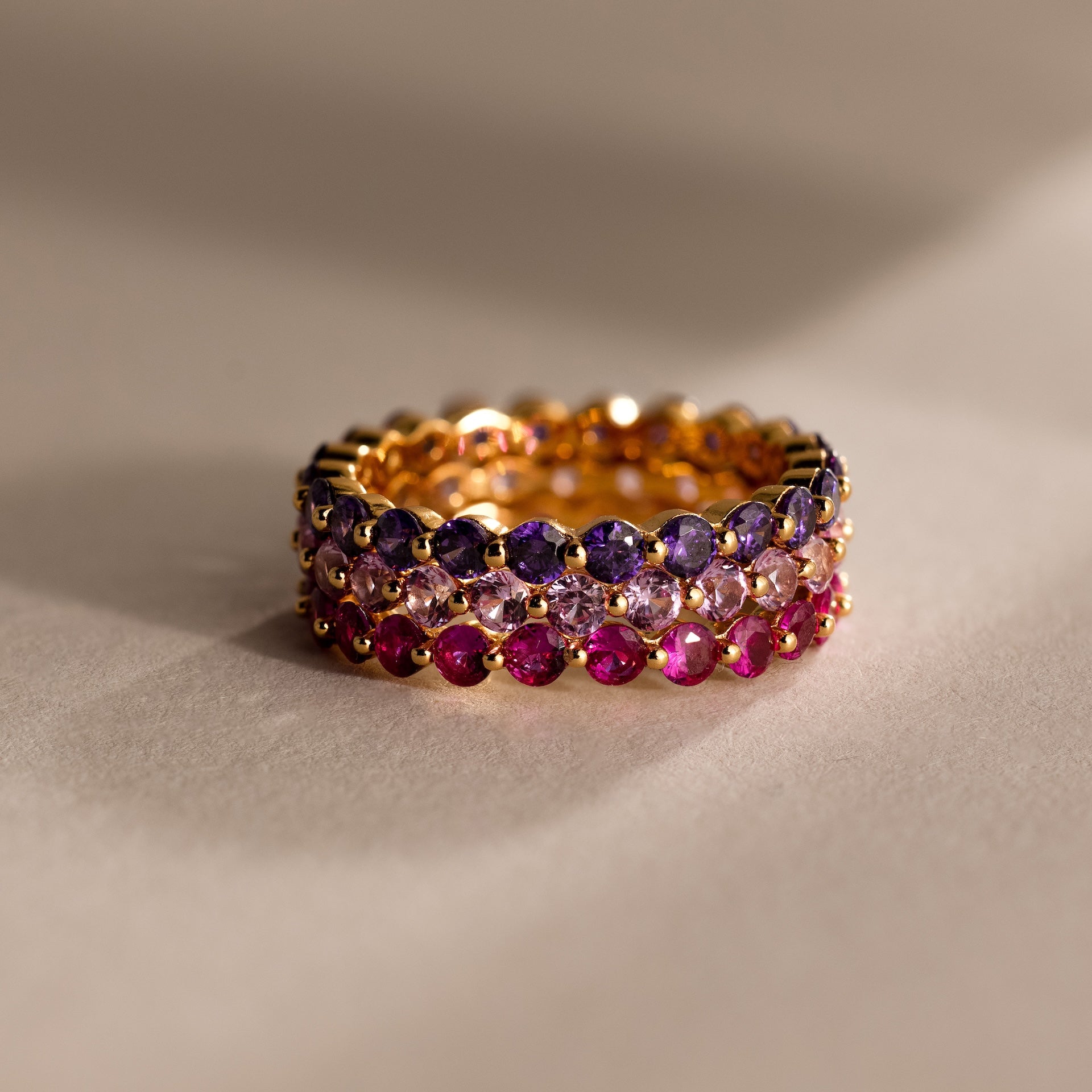 The Lace Eternity Ruby Ring, featuring three stacked gold bands with purple, clear, and pink gemstones, creates an elegant birthstone display against a neutral background.