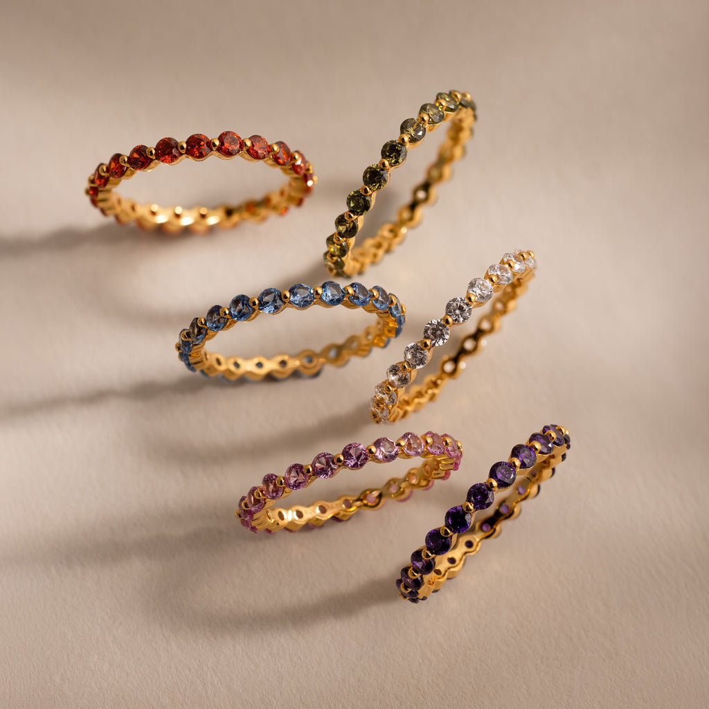 Six gold rings, including the Lace Eternity Garnet Ring with vibrant gemstones, are arranged in two rows on a beige surface.