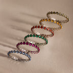 Six gemstone rings, featuring the Lace Eternity Emerald Ring, are arranged diagonally on a light beige surface with soft shadows.