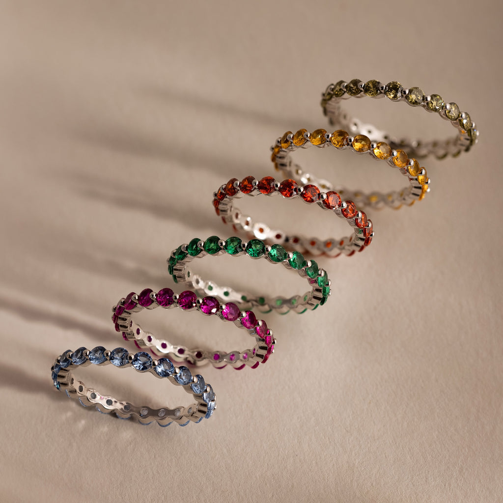 Six Lace Eternity Topaz Rings in vibrant blue, pink, green, red, orange, and yellow are arranged in a diagonal line on a beige surface.