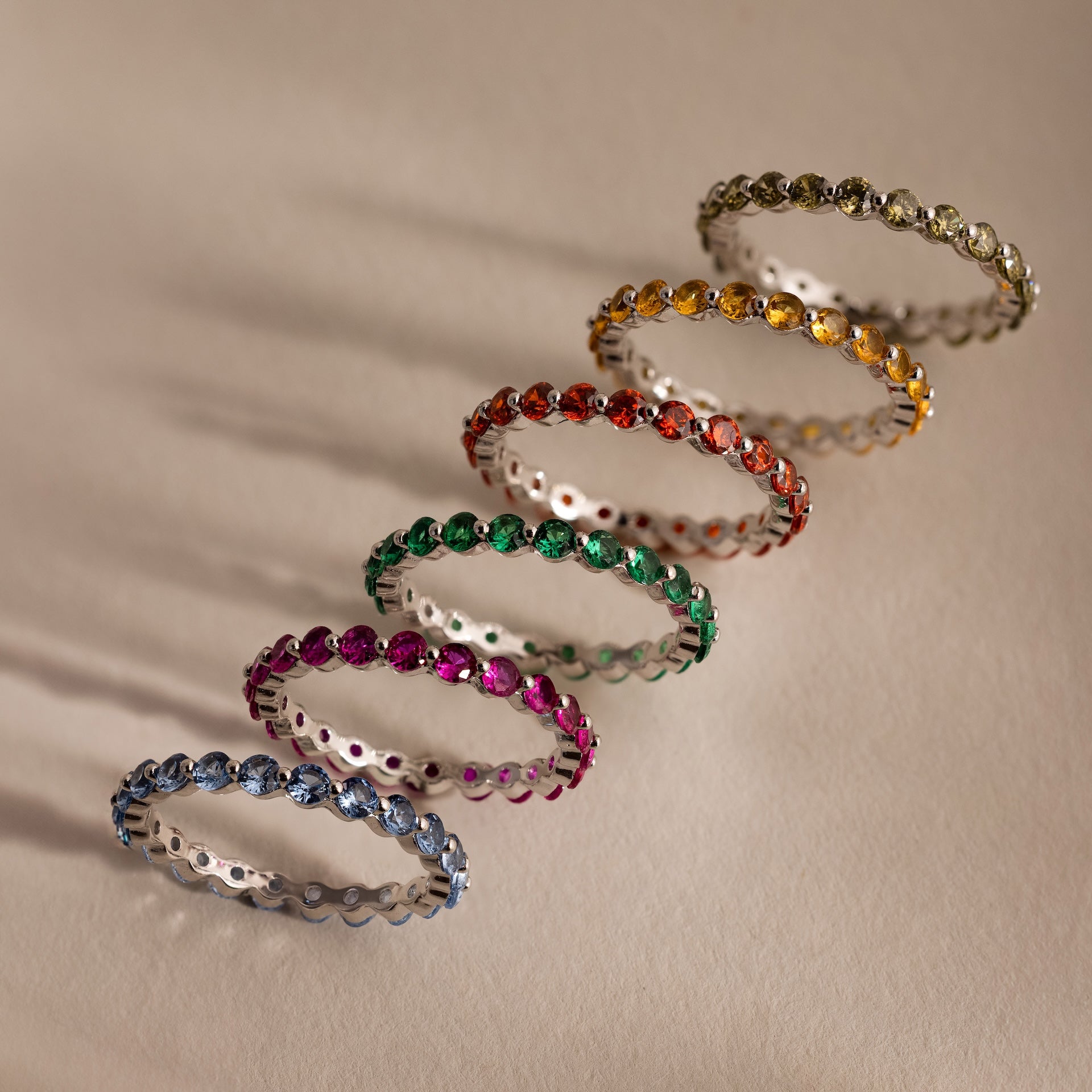 Six Lace Eternity Topaz Rings in vibrant blue, pink, green, red, orange, and yellow are arranged in a diagonal line on a beige surface.