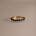 The Lace Eternity Sapphire Ring, a gold band adorned with small blue sapphires, is displayed on a neutral surface.