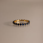 The Lace Eternity Sapphire Ring, a gold band adorned with small blue sapphires, is displayed on a neutral surface.