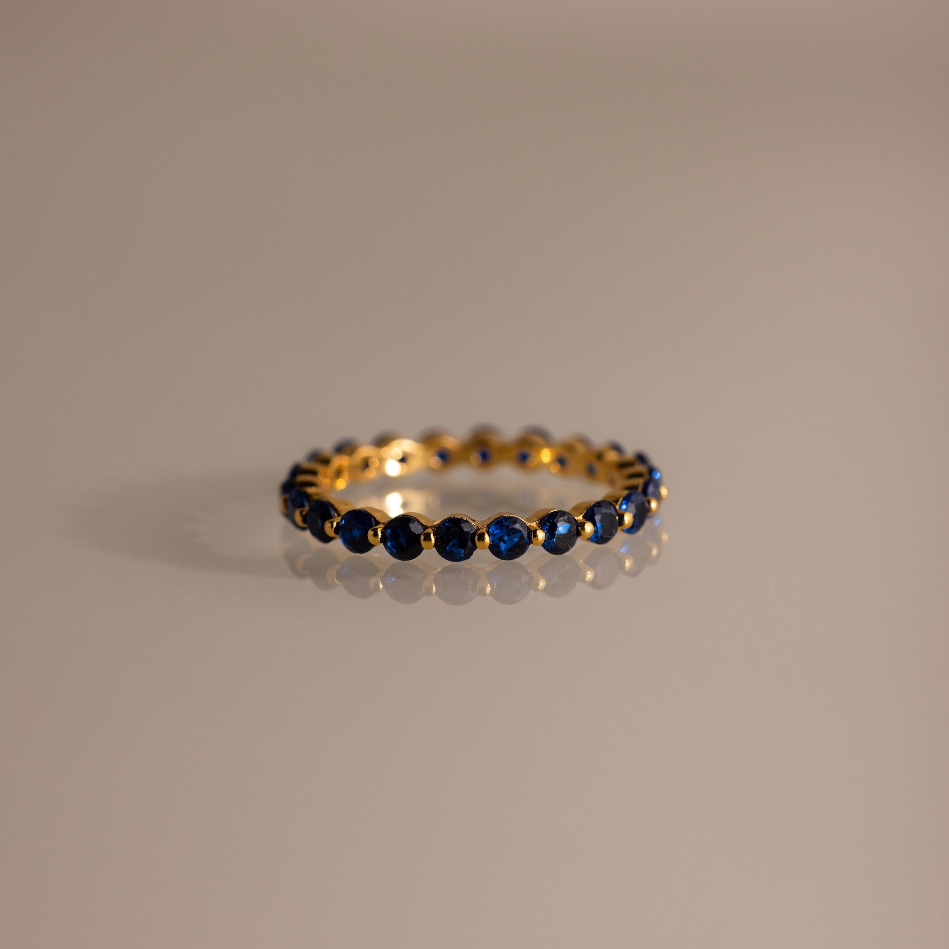 The Lace Eternity Sapphire Ring, a gold band adorned with small blue sapphires, is displayed on a neutral surface.