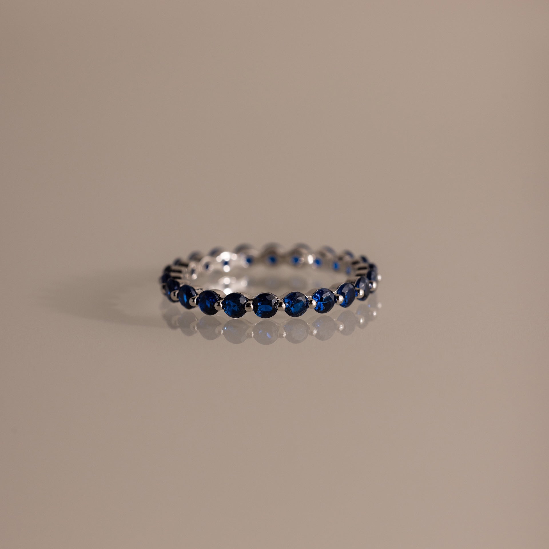 Lace Eternity Sapphire Ring displayed on a glossy light beige surface, featuring evenly spaced blue gemstones set in silver.
