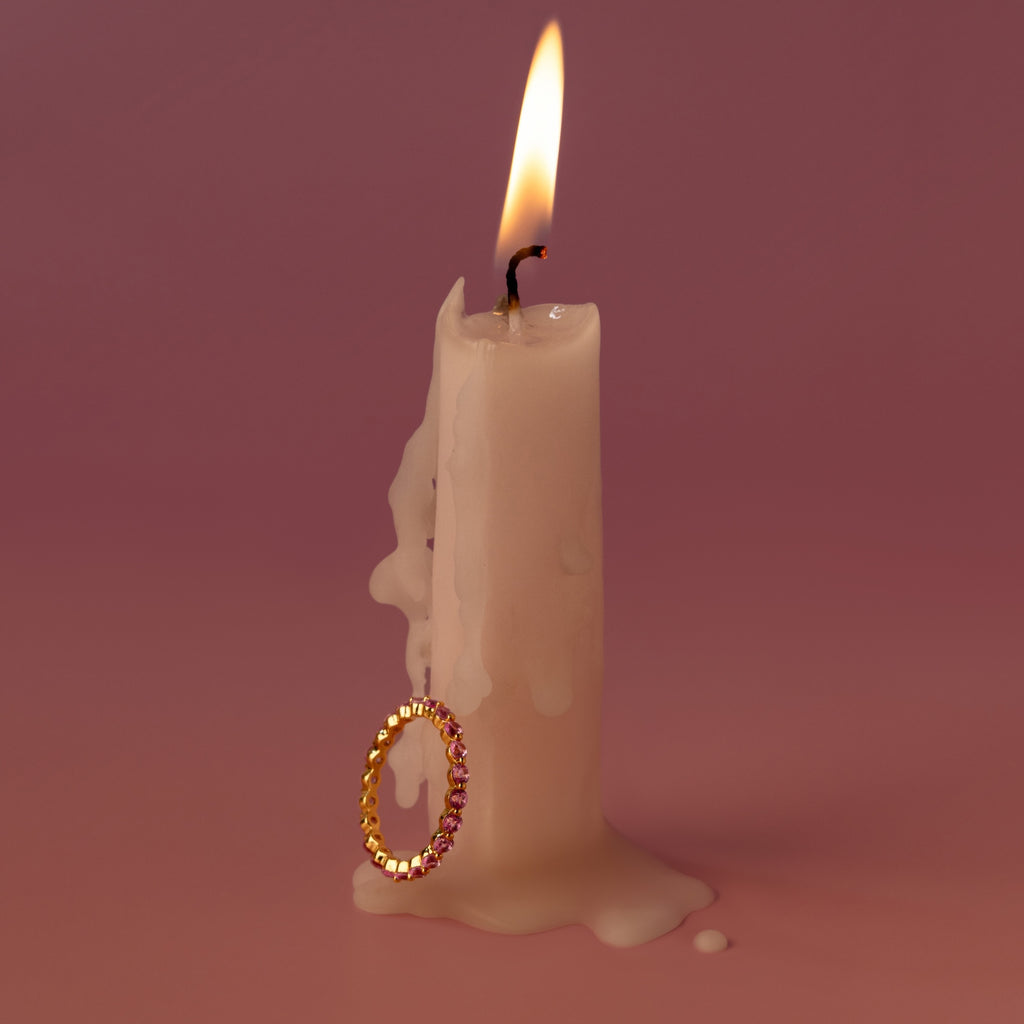 A white candle with melted wax is adorned with the Lace Eternity Pink Tourmaline Ring against a pink background.