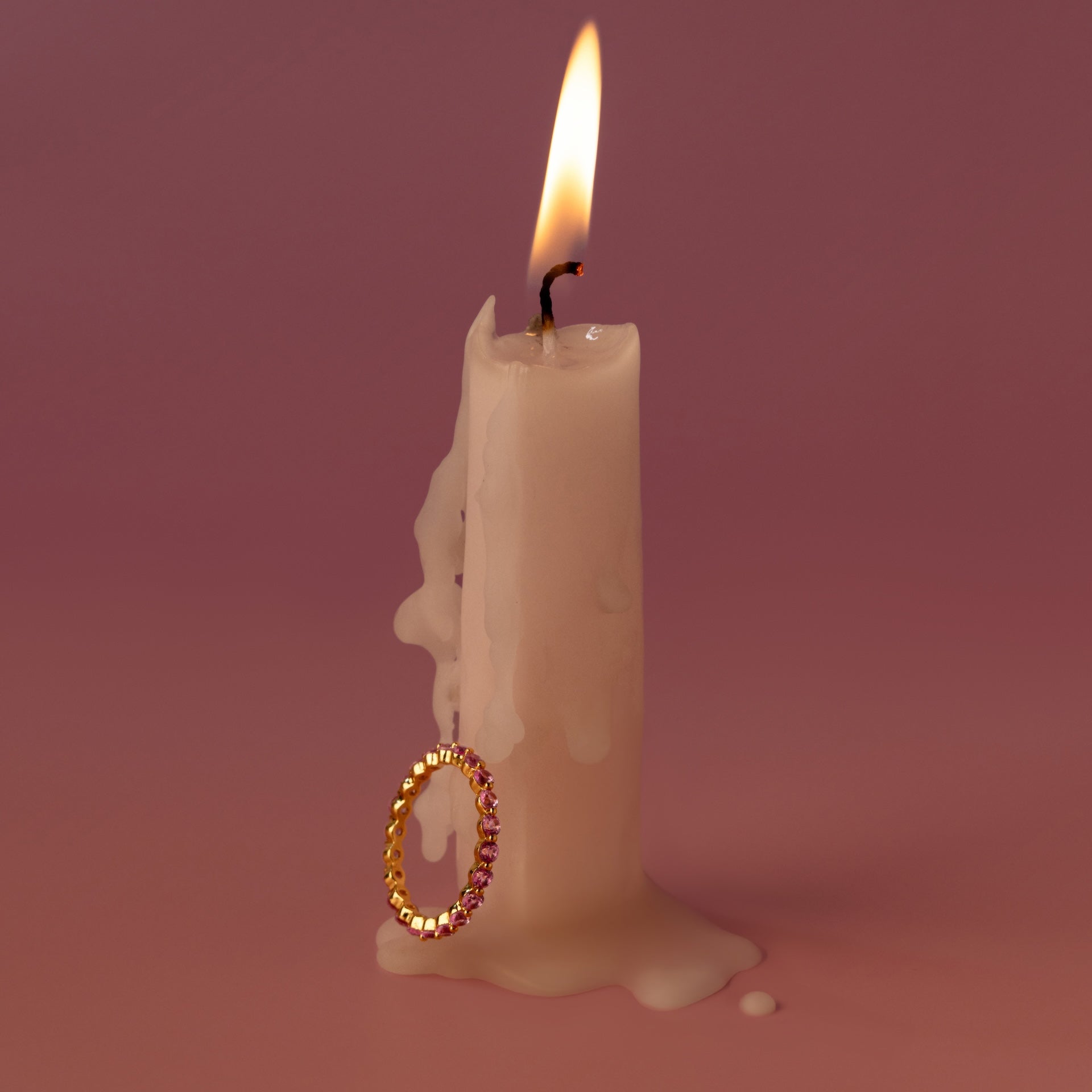 A white candle with melted wax is adorned with the Lace Eternity Pink Tourmaline Ring against a pink background.
