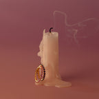 A melted candle with smoke rising and two gold rings, including a Lace Eternity Amethyst Ring, resting at its base on a pink background.