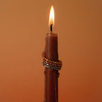 A lit brown candle with three jeweled rings, including the Lace Eternity Emerald Ring, stacked around it against a plain orange background.
