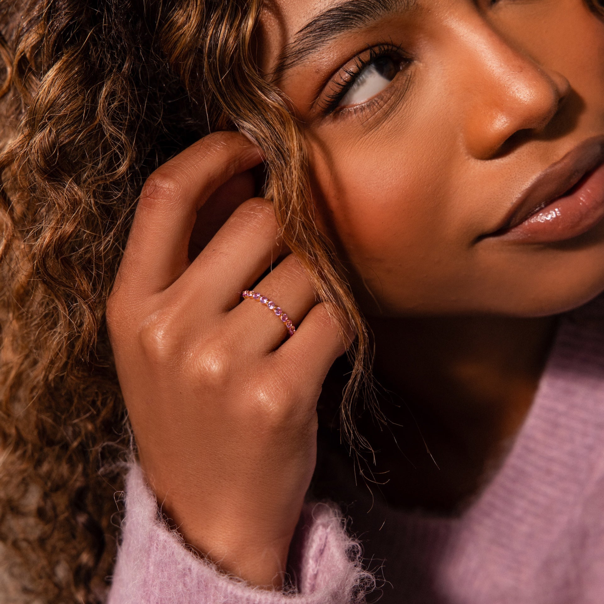 A woman with curly hair touches her face, wearing the Lace Eternity Birthstone Ring and a soft pink sweater with delicate lace accents.