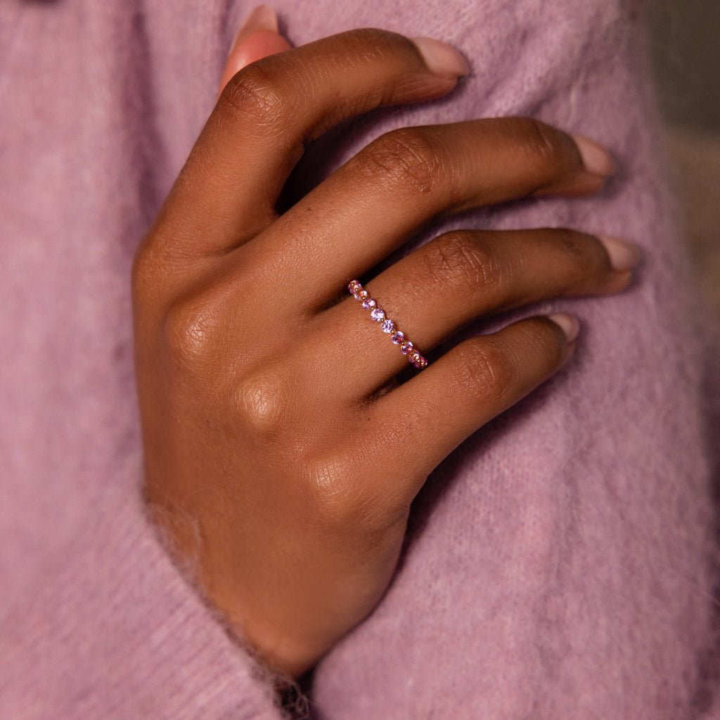 A hand wearing the Lace Eternity Alexandrite Ring with a pink birthstone rests on a lavender sweater, showcasing its delicate, lace-inspired design.