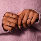 Two hands with manicured nails display multiple gold and gemstone rings, including the sparkling Lace Eternity Peridot Ring, against a soft pink sweater background.