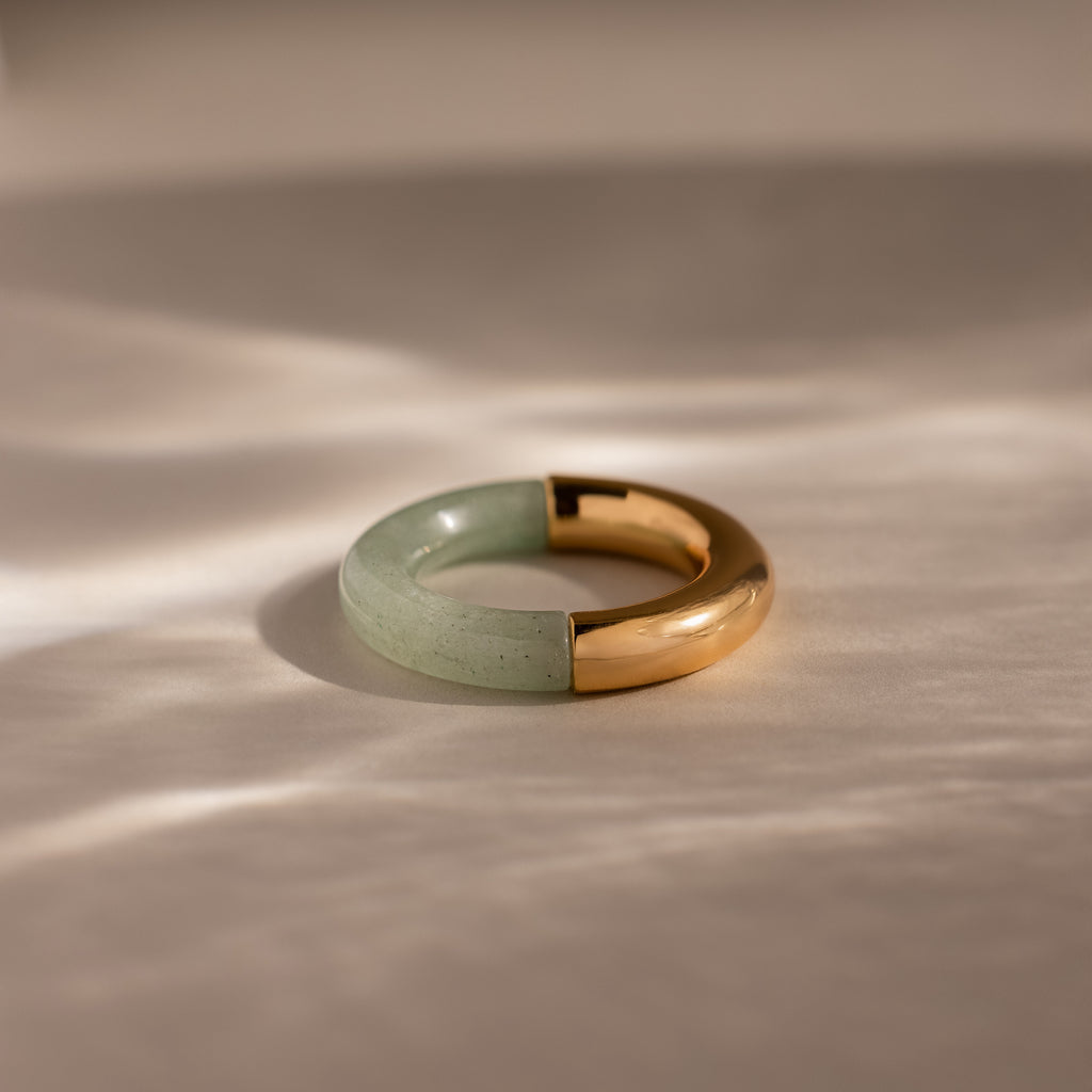 The Half Jade Ring is a round statement jewelry piece with a half-green jade stone and half-gold metal design, displayed on a light, textured surface.